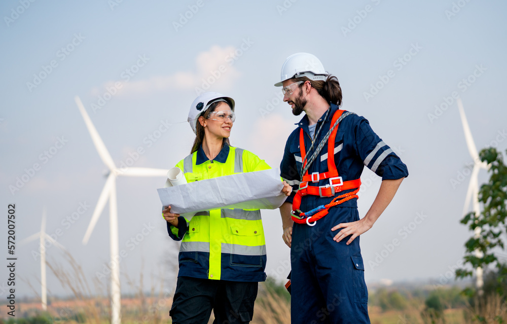 Caucasian engineer man discuss together with co-worker woman hold drawing paper and stay in front of row of windmill or wind turbine with blue sky. Caucasian engineer man discuss together with co-worker woman hold drawing paper and stay in front of row of windmill or wind turbine with blue sky.