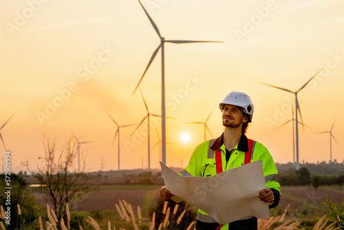 Caucasian engineer or technician man stand with holding drawiing paper and look to left side and stand in front of windmill or wind turbine cluster with sunset and warm light. photo