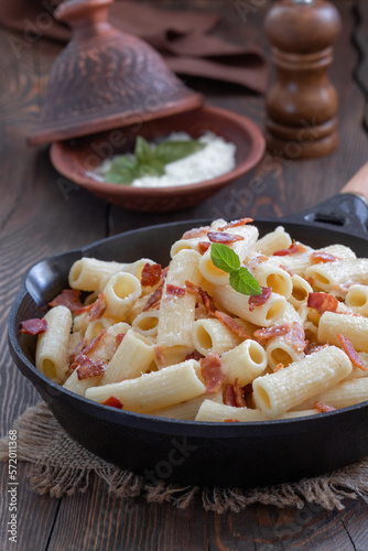 Pasta Alla Gricia. Rigatoni pasta with bacon and pecorino cheese served in frying pan with bowl of grated cheese on background. Dark wooden table, сlose-up, selective focus, vertical.