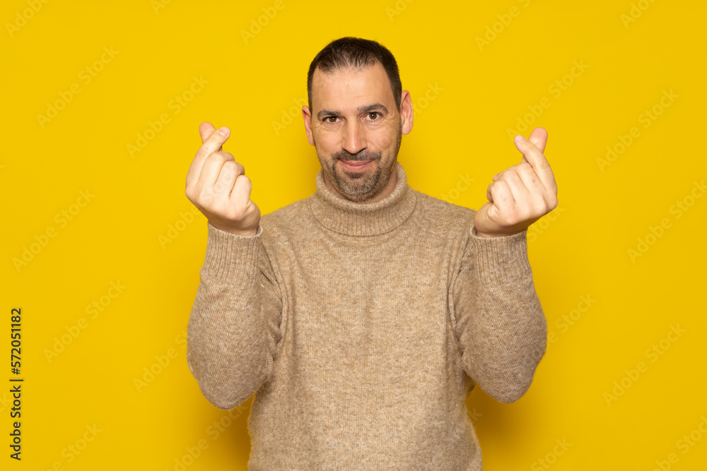Hispanic man with a beard wearing a turtleneck making the money gesture with his fingers isolated over yellow background. Hispanic man with a beard wearing a turtleneck making the money gesture with his fingers isolated over yellow background.
