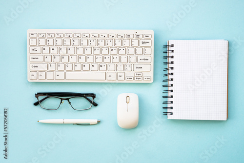 Office workspace with keyboard, notepad, glasses, pen and coffee cup. Flat lay image on blue, minimal.