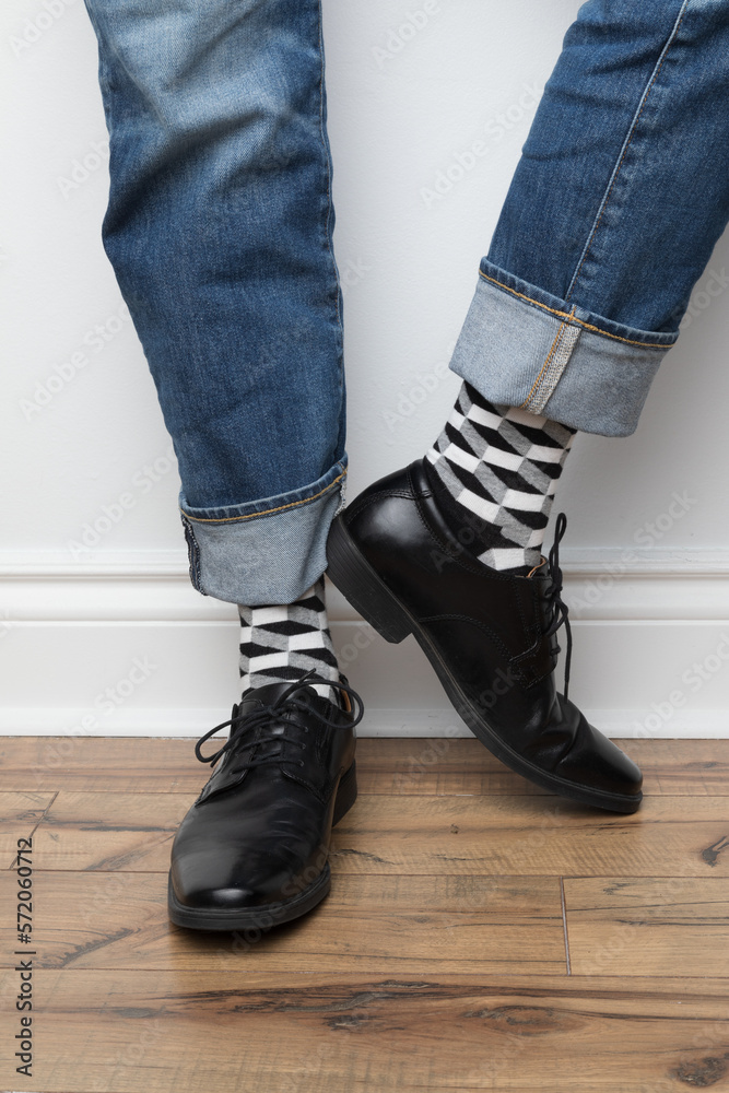 Product photos of men's footwear. Close up photo of a man's foot and legs. He is wearing blue jeans, black and white patterned socks and black dress shoes.