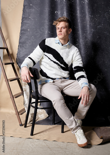 a stylish man in a fashionable sweater poses sitting in the studio