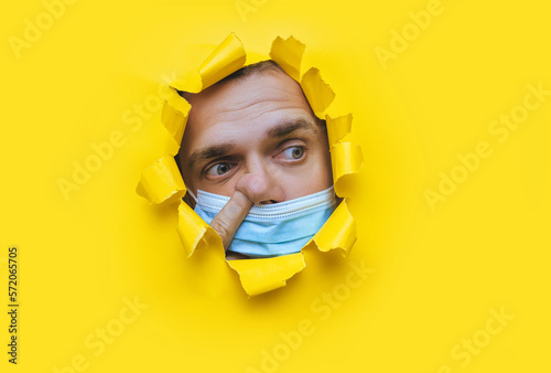 A man in a blue medical mask looks in surprise into a broken hole in yellow paper and picks his nose with his finger. The concept of pandemic, quarantine, protection against coronavirus and covid-19.