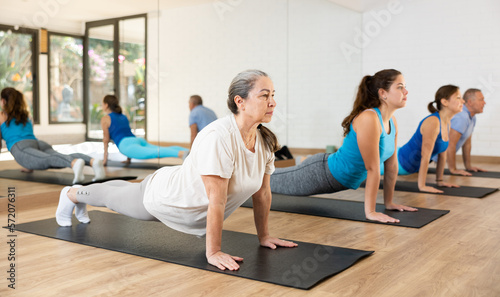 Willing aged woman engaging in pilates training on mat in gym room during workout session. Persons practicing pilates in fitness studio