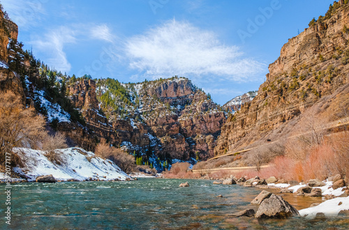 Beautiful view of river floating through canyon in winter; fast running water in foreground; mountains and blue sky in background; distant view of highway on the right