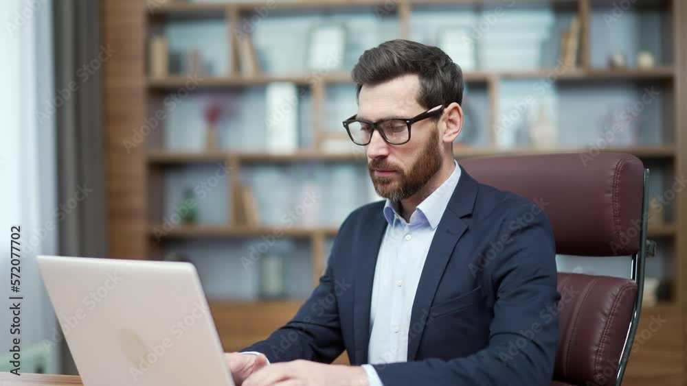 Busy mature business man investor with glasses typing on laptop computer working online at desktop in modern office Concentrated bearded entrepreneur takes responsible approach to the project indoors
