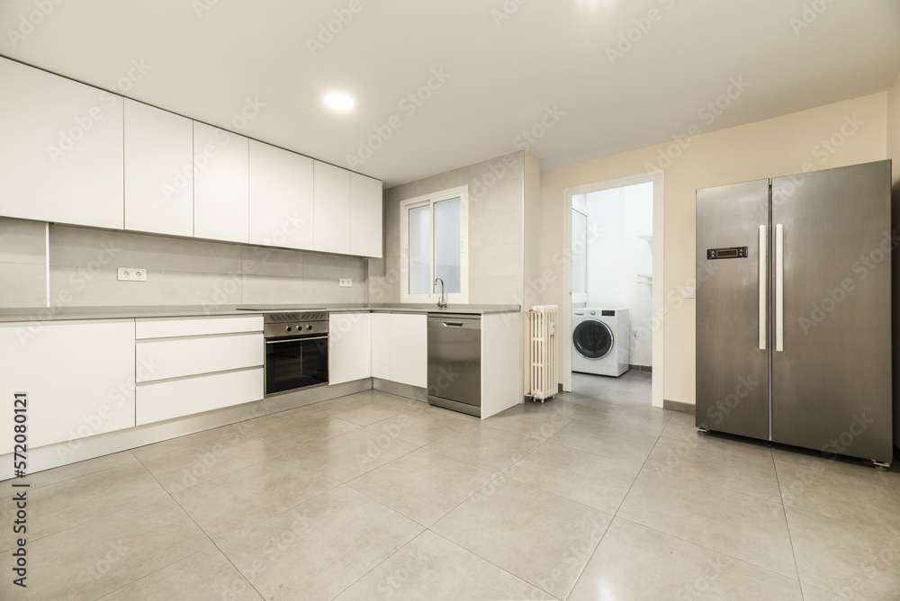 Large kitchen with modern furniture without handles with white doors and drawers, stoneware floors with large gray tiles, appliances and a large stainless steel refrigerator