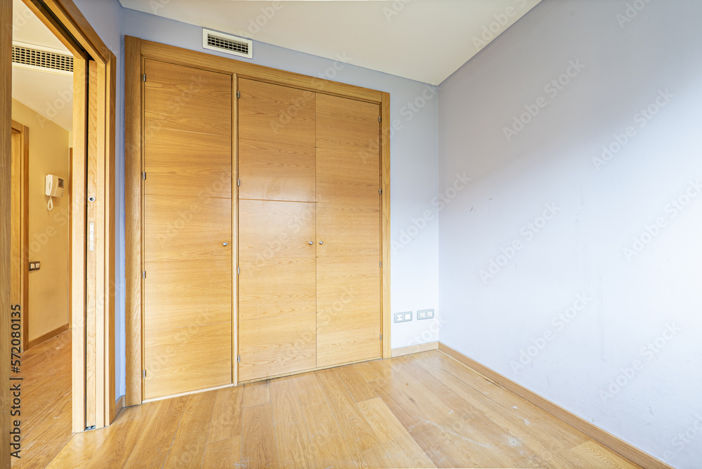 Empty room with a built-in wardrobe with three folding wooden doors, smooth blue walls and laminated oak flooring