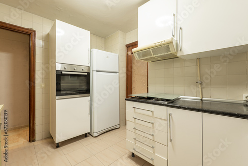 Corner of an old kitchen with white furniture, a column with an oven and a white fridge