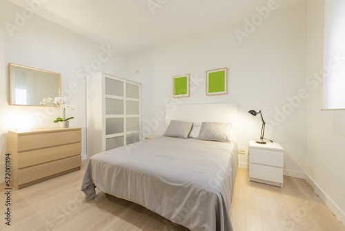 Double bedroom with white wooden headboard, gray bedspread and cushions, white bedside table with folding lamp and oak chest of drawers with matching mirror