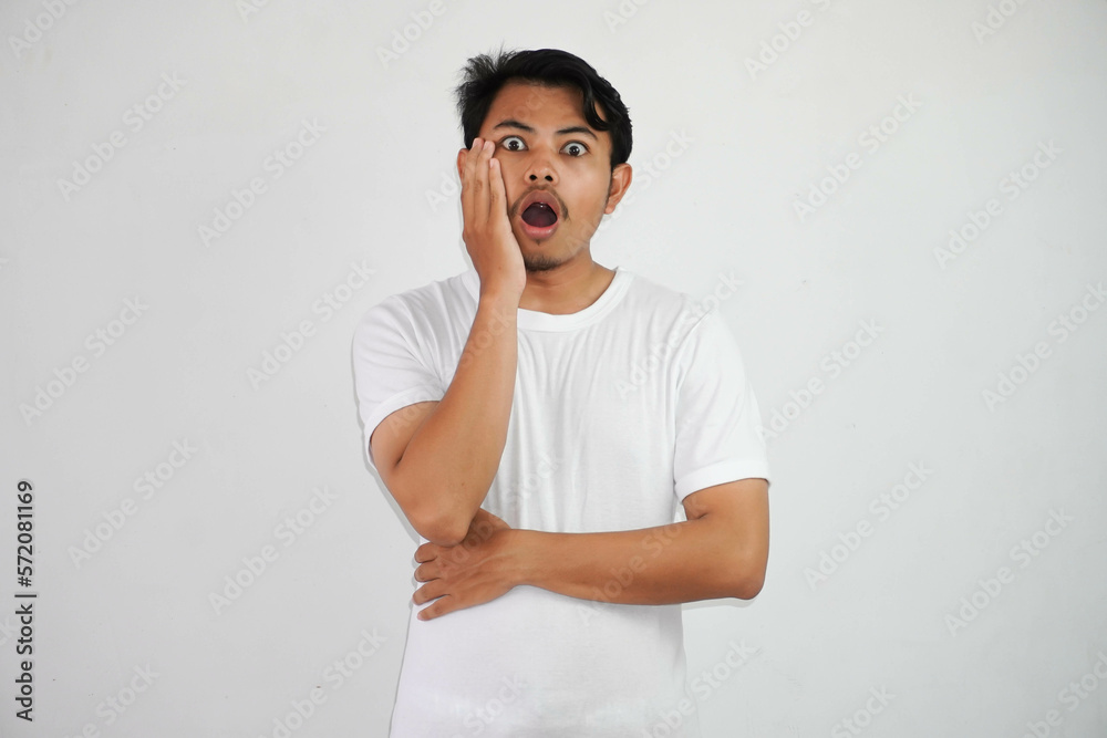 Shocked young Asian man standing hand on cheek with open mouth wearing white t shirt isolated on white background Shocked young Asian man standing hand on cheek with open mouth wearing white t shirt isolated on white background