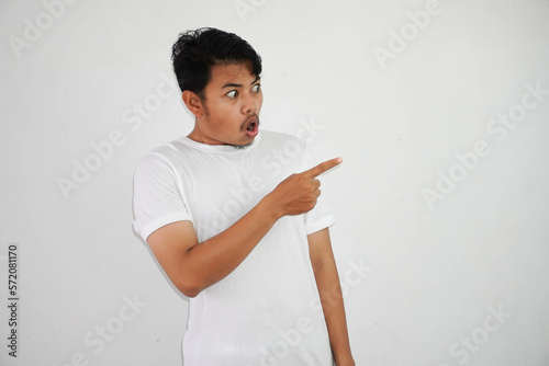 shocked asian man with fingers pointing to the side wearing white t shirt isolated on white background