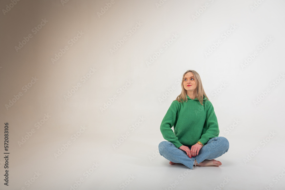 Young, cute, emotional, chic girl sitting on the floor in a flirtatious mood, hand gestures, empty space, wearing jeans and a green sweater on an isolated white background, place for text