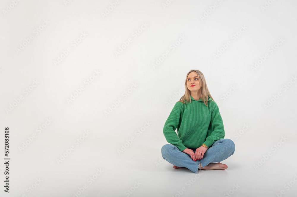 Young, cute, emotional, chic girl sitting on the floor in a flirtatious mood, hand gestures, empty space, wearing jeans and a green sweater on an isolated white background, place for text Young, cute, emotional, chic girl sitting on the floor in a flirtatious mood, hand gestures, empty space, wearing jeans and a green sweater on an isolated white background, place for text