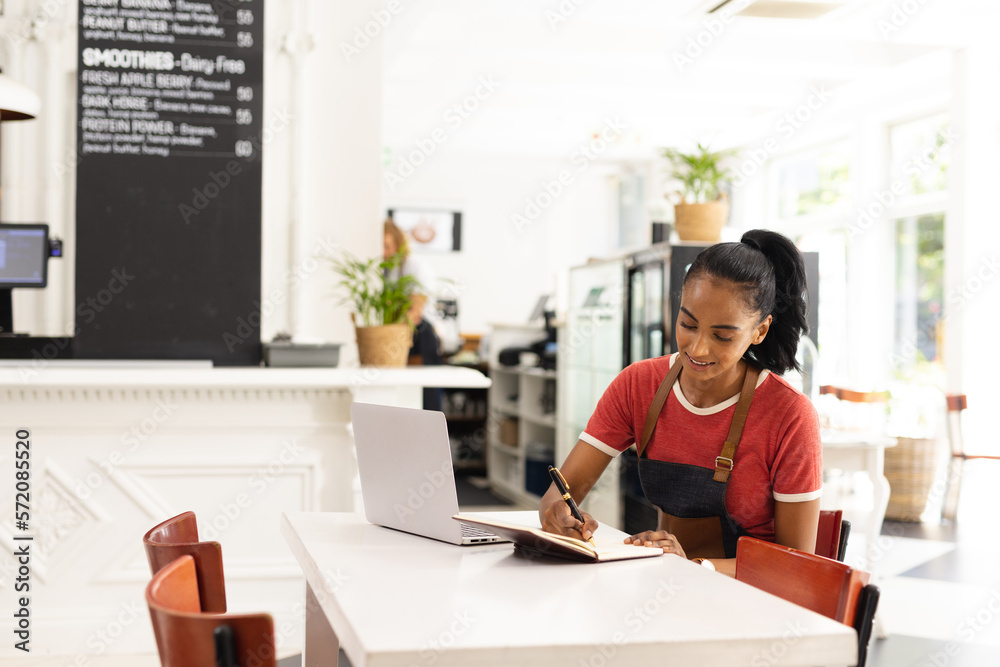 Happy biracial female barista wearing apron and taking notes in cafe Happy biracial female barista wearing apron and taking notes in cafe