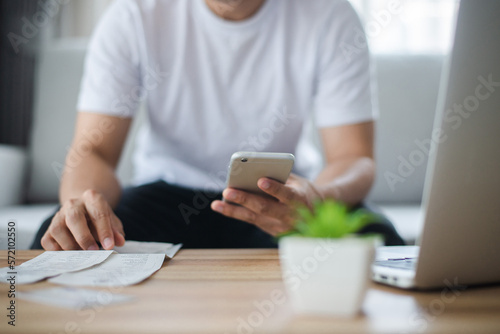 Young man wearing wear casual clothes hand holding and using smart phone paying bills related to home expenses such as water and electricity with calculator and laptop on wooden desk at home.