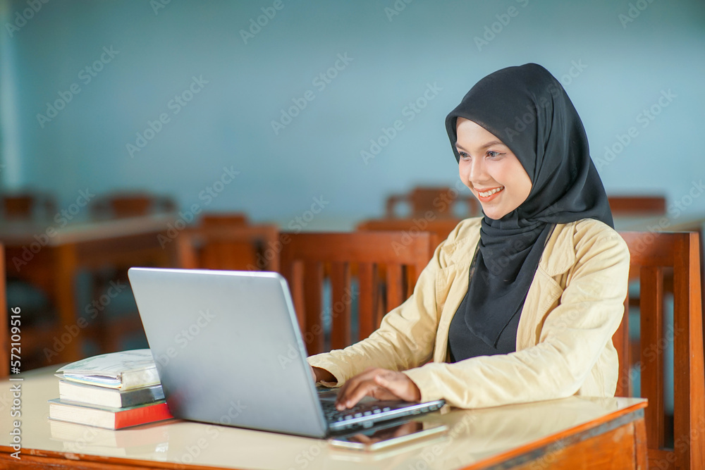 beautiful asian young woman wearing hijab smiling at work, typing operating laptop computer and mobile phone at her desk. beautiful asian young woman wearing hijab smiling at work, typing operating laptop computer and mobile phone at her desk.