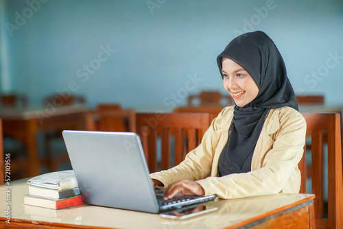 beautiful asian young woman wearing hijab smiling at work, typing operating laptop computer and mobile phone at her desk.