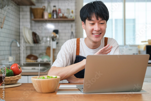 Smiling man having breakfast in modern kitchen at home and making video call via laptop