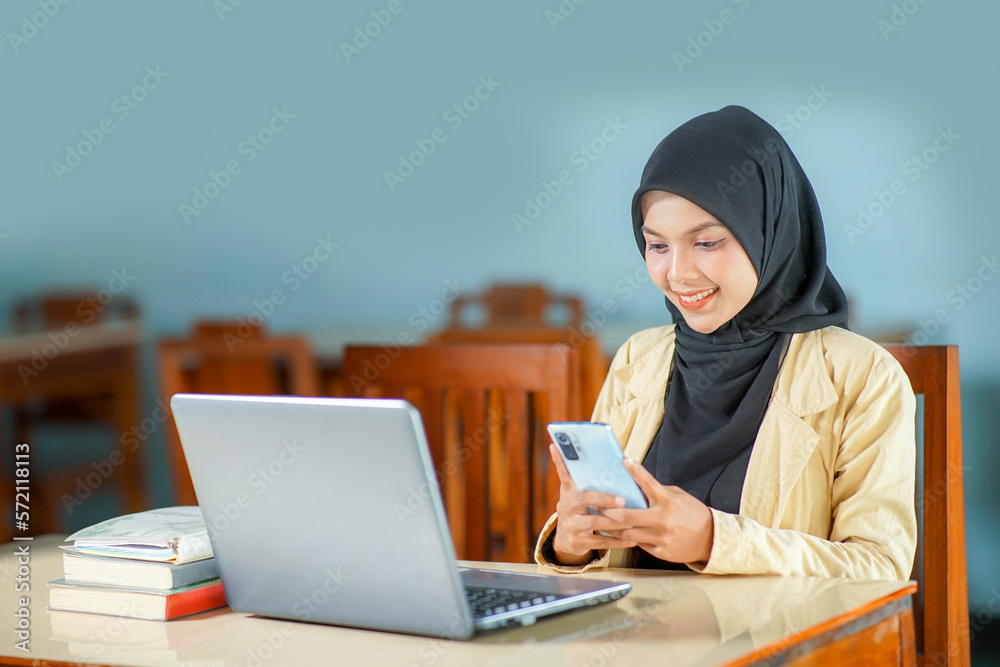 beautiful young asian woman wearing hijab smiling at work, operating a mobile phone and laptop computer at her desk. beautiful young asian woman wearing hijab smiling at work, operating a mobile phone and laptop computer at her desk.