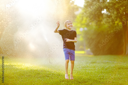 Funny little boy playing with garden sprinkler in sunny city park. Elementary school child laughing, jumping and having fun with spray of water. Summer outdoors activity for kids.