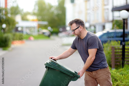 Mature man pulling out a large green plastic garbage container in front of the townhouse to the roadway of the street. Garbage collection and recycling trash photo