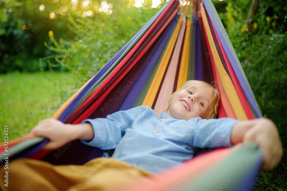 Cute little blond white boy enjoy and having fun with multicolored hammock in backyard or outdoor playground. Summer outdoors active leisure for kids. Child relaxing and swinging in hammock. Cute little blond white boy enjoy and having fun with multicolored hammock in backyard or outdoor playground. Summer outdoors active leisure for kids. Child relaxing and swinging in hammock.