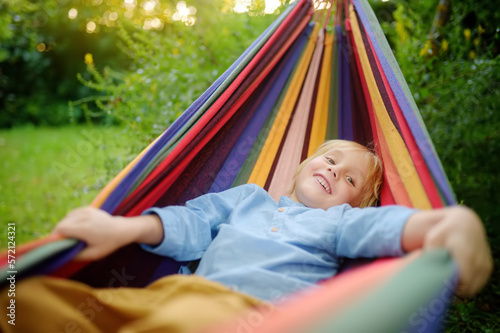 Cute little blond white boy enjoy and having fun with multicolored hammock in backyard or outdoor playground. Summer outdoors active leisure for kids. Child relaxing and swinging in hammock.