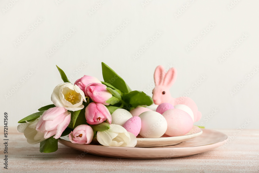Plate with Easter eggs, tulip flowers and bunny on table near light wall