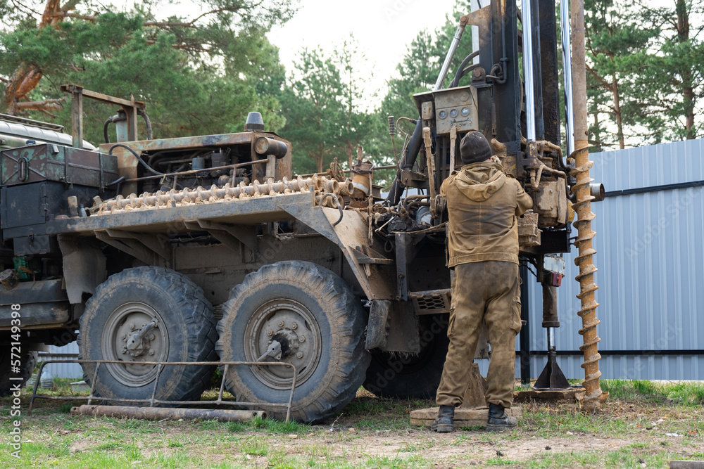 Obraz premium Team of workers with drilling rig on car are drilling artesian well for water in ground. Insertion of metal casing pipe into ground, installation of individual drinking supply, June 28, 2022, Russia,