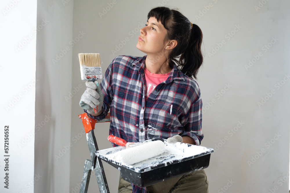 puzzled woman in paint roller and white paint for walls in hands close-up portrait. Construction work and cosmetic repairs in house, wall painting, tinting, finishing work with your own hands