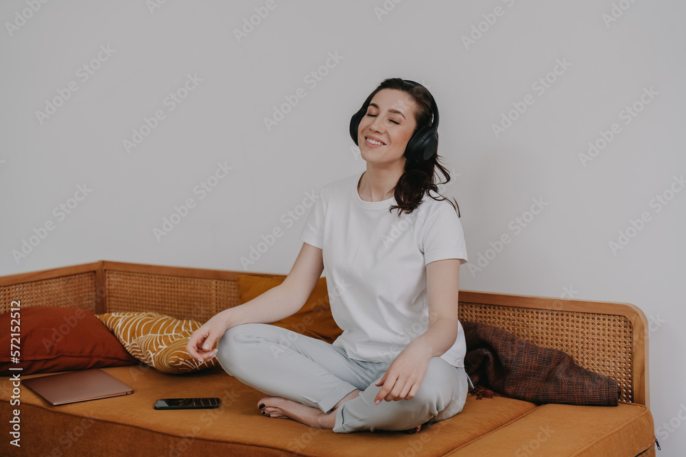 Indoor shoot of  meditating beautiful woman sitting on orange sofa at home legs crossed at home with headphones. Girl relaxing after studies, peacefully sitting in a  yoga posture. Calm housewife
