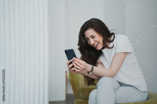 Laughing brunette young woman in casual sits on chair holds phone looks at screen in disbelief reads message smiles. Happy female relaxing home on vacation. Domestic leisure, people sincere emotions.