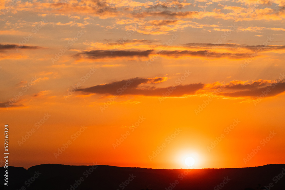 Summer sunset with yellow and orange colours. Beautiful clouds in the sky. The sun hovers over the mountain. The sun's rays from below illuminate the dark clouds. The beauty of nature.