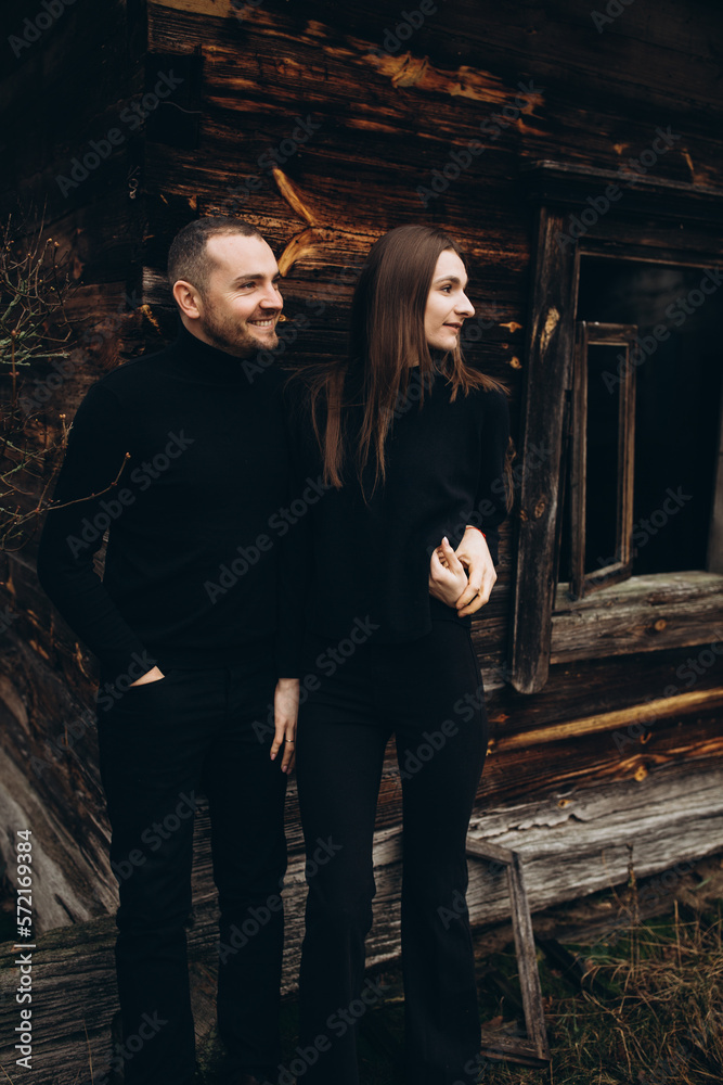 a man and a woman in black clothes near an old wooden house. photo in dark brown tones. old abandoned house and cloudy cold weather. beautiful stylish couple on the background of a brown wooden wall.
