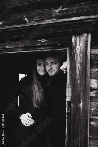 a man and a woman in black clothes near an old wooden house. photo in dark brown tones. old abandoned house and cloudy cold weather. beautiful stylish couple on the background of a brown wooden wall. © Anhelina Tyshkovets
