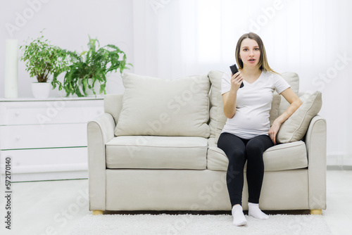 Pregnant woman sitting on sofa with remote control.
