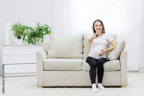 Cute pregnant woman sitting on sofa with remote control.