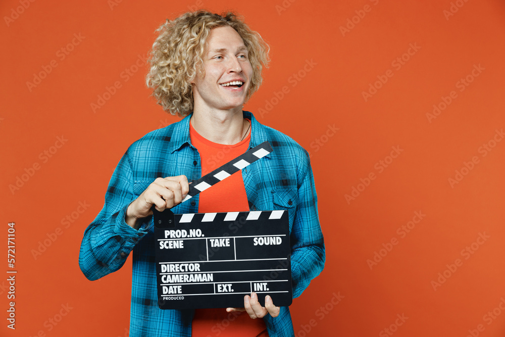 Young blond caucasian man wear blue shirt orange t-shirt hold in hand classic black film making clapperboard look aside area isolated on plain red background studio portrait. People lifestyle concept.
