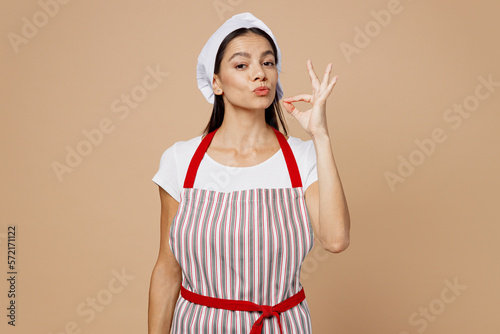 Young housewife housekeeper chef cook baker latin woman in striped apron toque hat making okay taste delight sign, savoring, delicious isolated on plain pastel beige background. Cooking food concept.