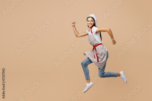 Full body side view smiling happy fun young housewife housekeeper chef baker latin woman wear apron toque hat run fast look camera isolated on plain pastel light beige background. Cook food concept.