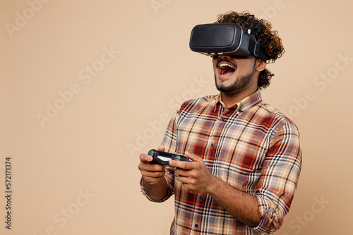 Young fun Indian man wear brown shirt casual clothes hold in hand play pc game with joystick console watching in vr headset pc gadget isolated on plain pastel light beige background studio portrait.