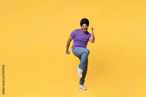 Full body fun devotee Sikh Indian man ties his traditional turban dastar wear purple t-shirt doing winner gesture celebrate clenching fists say yes isolated on plain yellow background studio portrait.