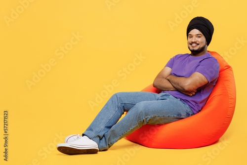 Obraz na plátně Full body fun devotee Sikh Indian man ties his traditional turban dastar wear purple t-shirt sit in bag chair hold hands crossed folded look camera isolated on plain yellow background studio portrait