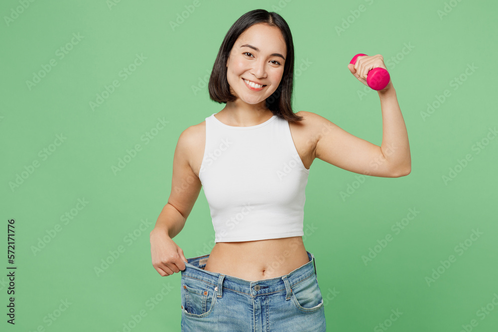 Young woman wears white clothes show loose pants on waist after weightloss hold dumbbell isolated on plain pastel light green background. Proper nutrition healthy fast food unhealthy choice concept. Young woman wears white clothes show loose pants on waist after weightloss hold dumbbell isolated on plain pastel light green background. Proper nutrition healthy fast food unhealthy choice concept.