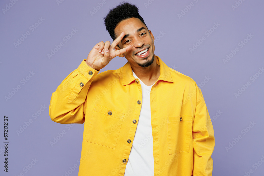 Young happy man of African American ethnicity wear yellow shirt t-shirt showing cover eye with victory sign isolated on plain pastel light purple background studio portrait. People lifestyle concept. Young happy man of African American ethnicity wear yellow shirt t-shirt showing cover eye with victory sign isolated on plain pastel light purple background studio portrait. People lifestyle concept.