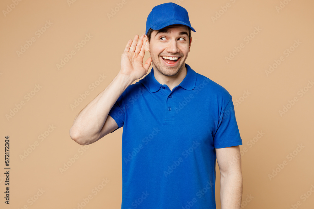 Professional delivery guy employee man wear blue cap t-shirt uniform workwear work as dealer courier try hear you overhear listening intently isolated on plain light beige background. Service concept.