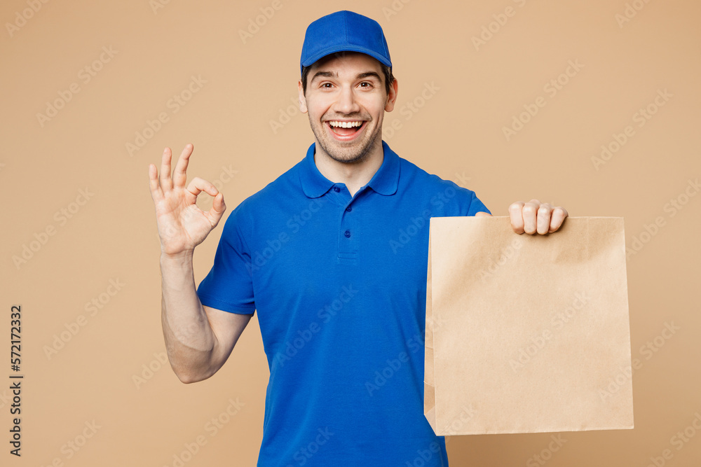Delivery guy employee man wear blue cap t-shirt uniform workwear work as dealer courier hold brown clear blank craft paper takeaway bag mock up show ok okay isolated on plain light beige background.