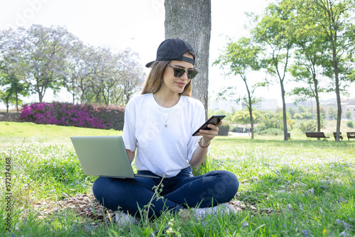 Front view of a happy student girl working with a laptop and smartphone in a green park of an university campus photo